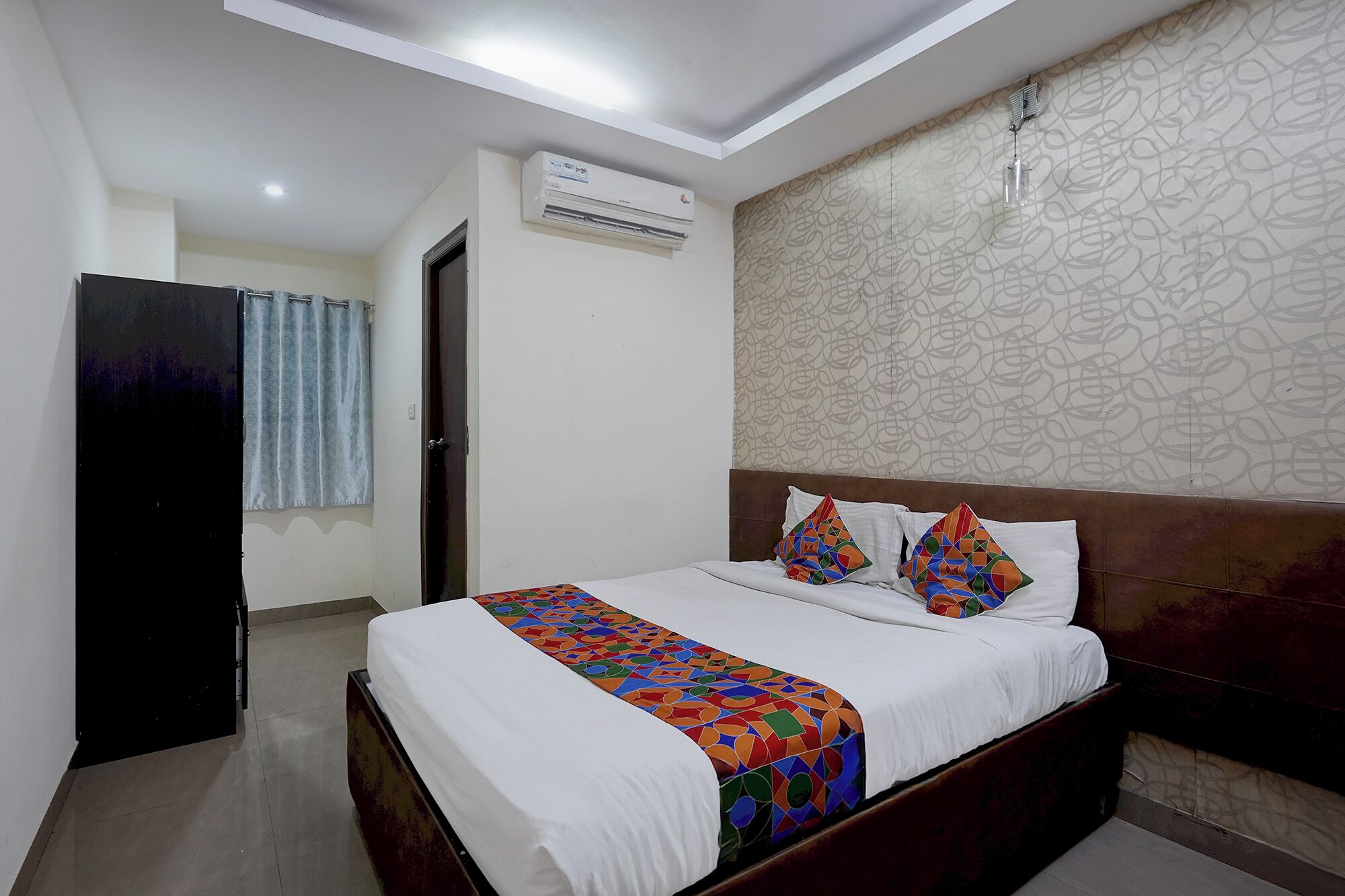 A hotel room with a bed, a window with curtains, an air conditioning unit, and a patterned wall.