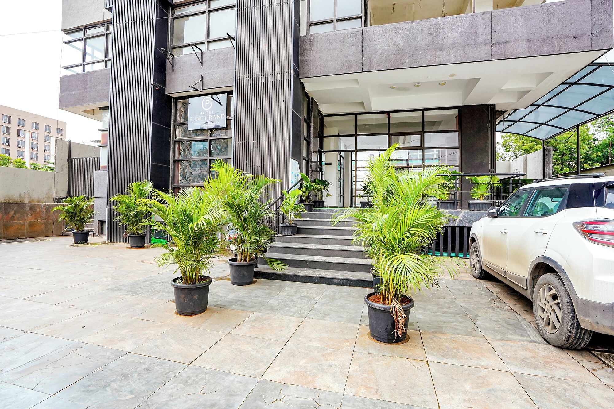 A hotel entrance with a modern design, featuring a staircase leading to the entrance, potted plants, and a parked car.