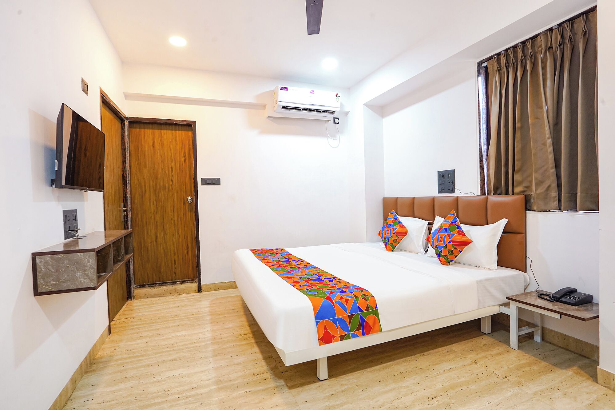 Habitación de hotel con cama, mesita de noche, televisor, aire acondicionado y ventana con cortinas.
