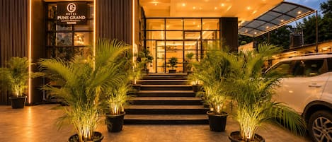 Entrada de un hotel con un letrero que dice 'Hotel Pune Grand Rooms', una escalera que conduce a la entrada y plantas en macetas en el suelo.