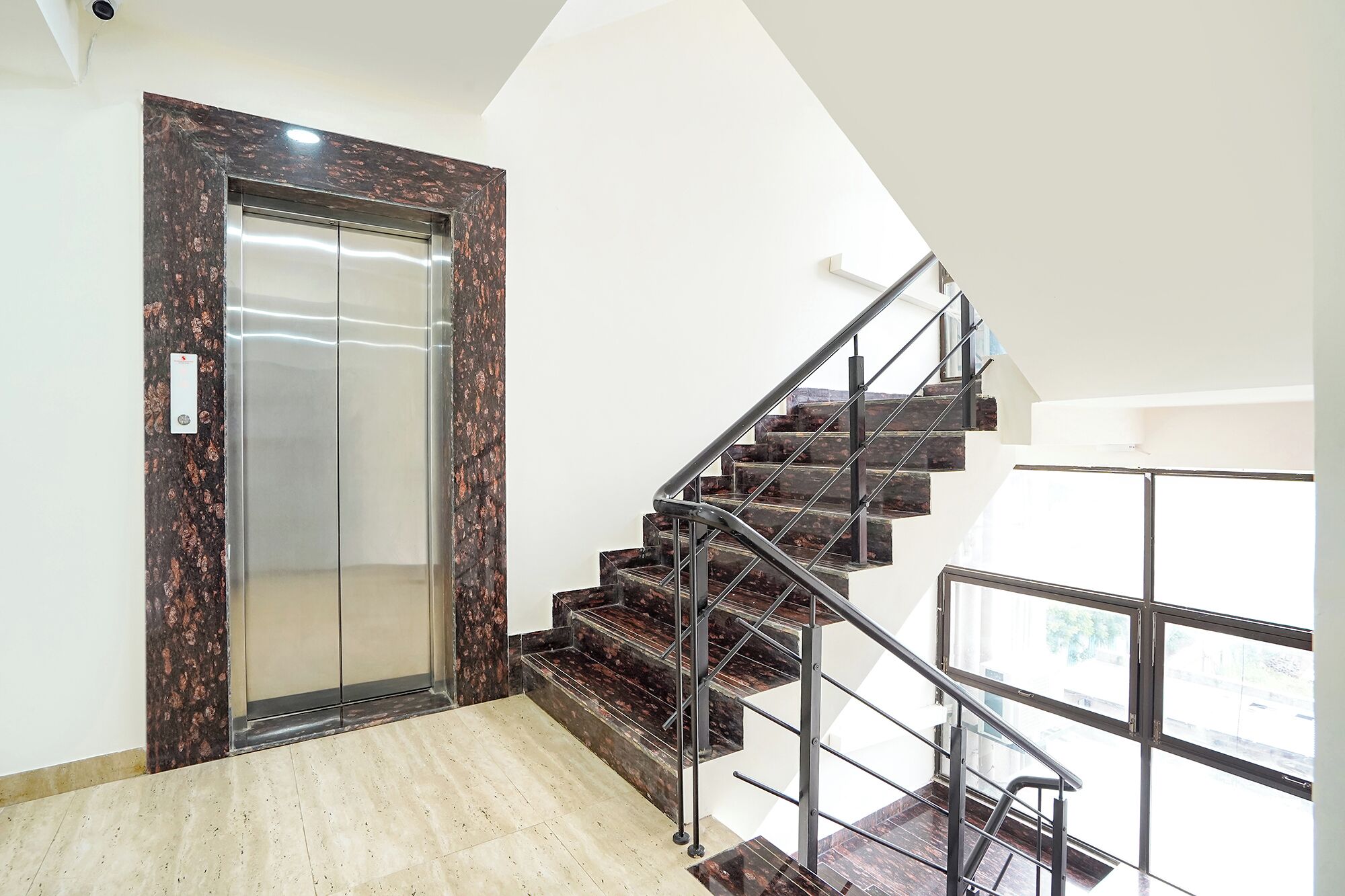 A modern interior with a marble elevator, a staircase with metal railings, and a large window providing natural light.