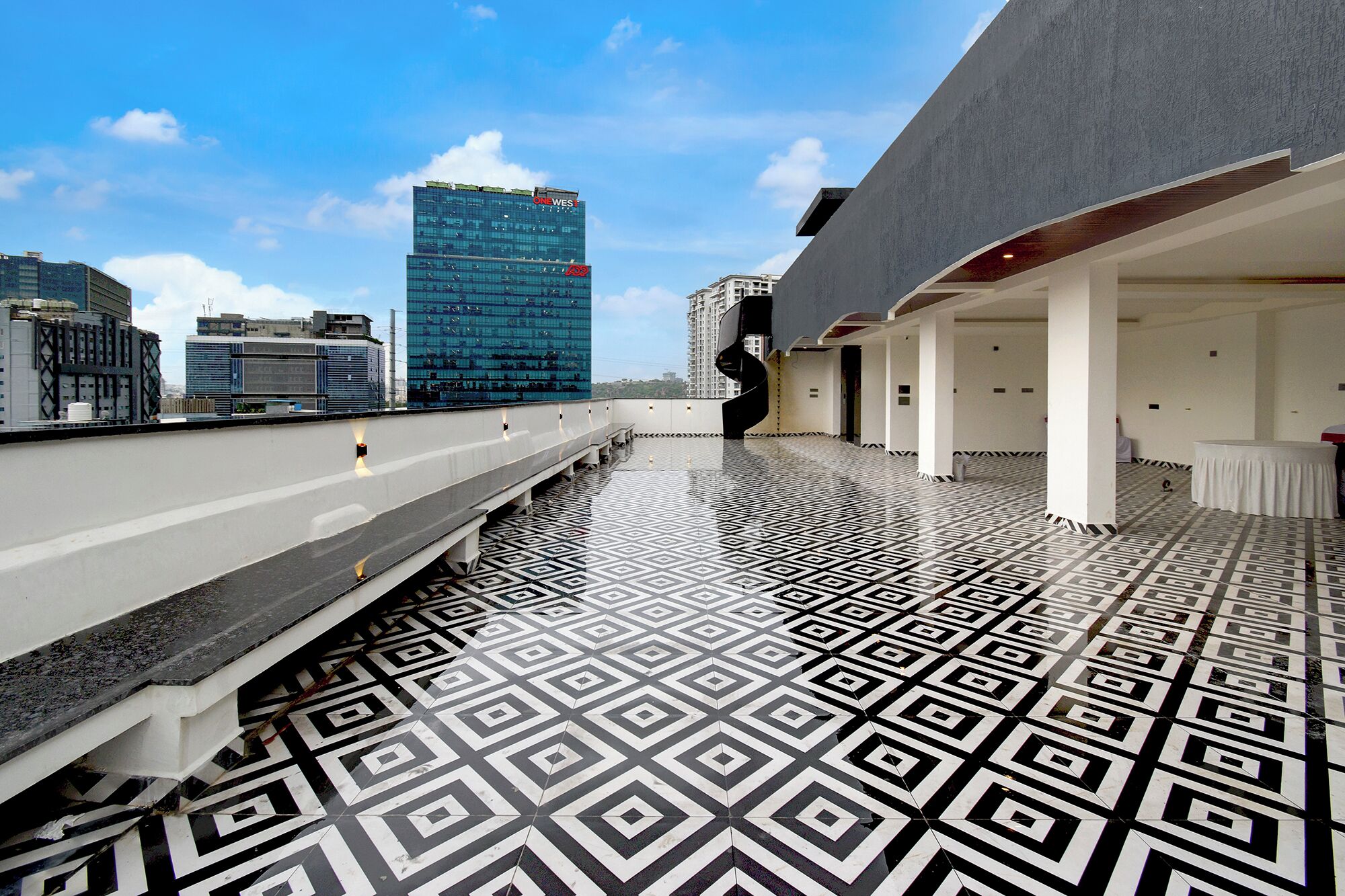 Een terras op het dak met een vloer met een geometrisch patroon, een modern gebouw met een glazen gevel en een heldere lucht.
