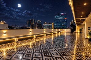 A rooftop terrace with patterned flooring, a spiral staircase, and a city skyline with illuminated buildings in the background.