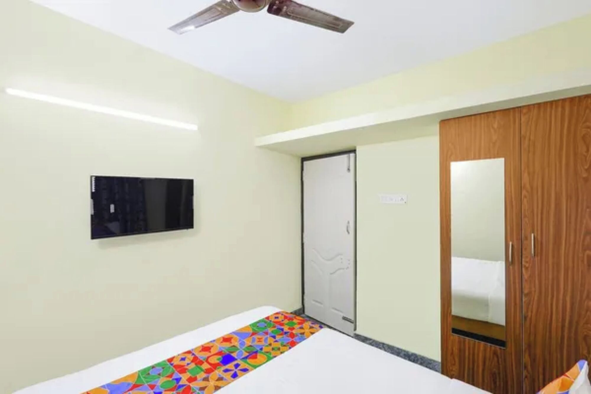 Une chambre à coucher comprenant un lit, un ventilateur de plafond, un téléviseur fixé au mur et une armoire en bois.
