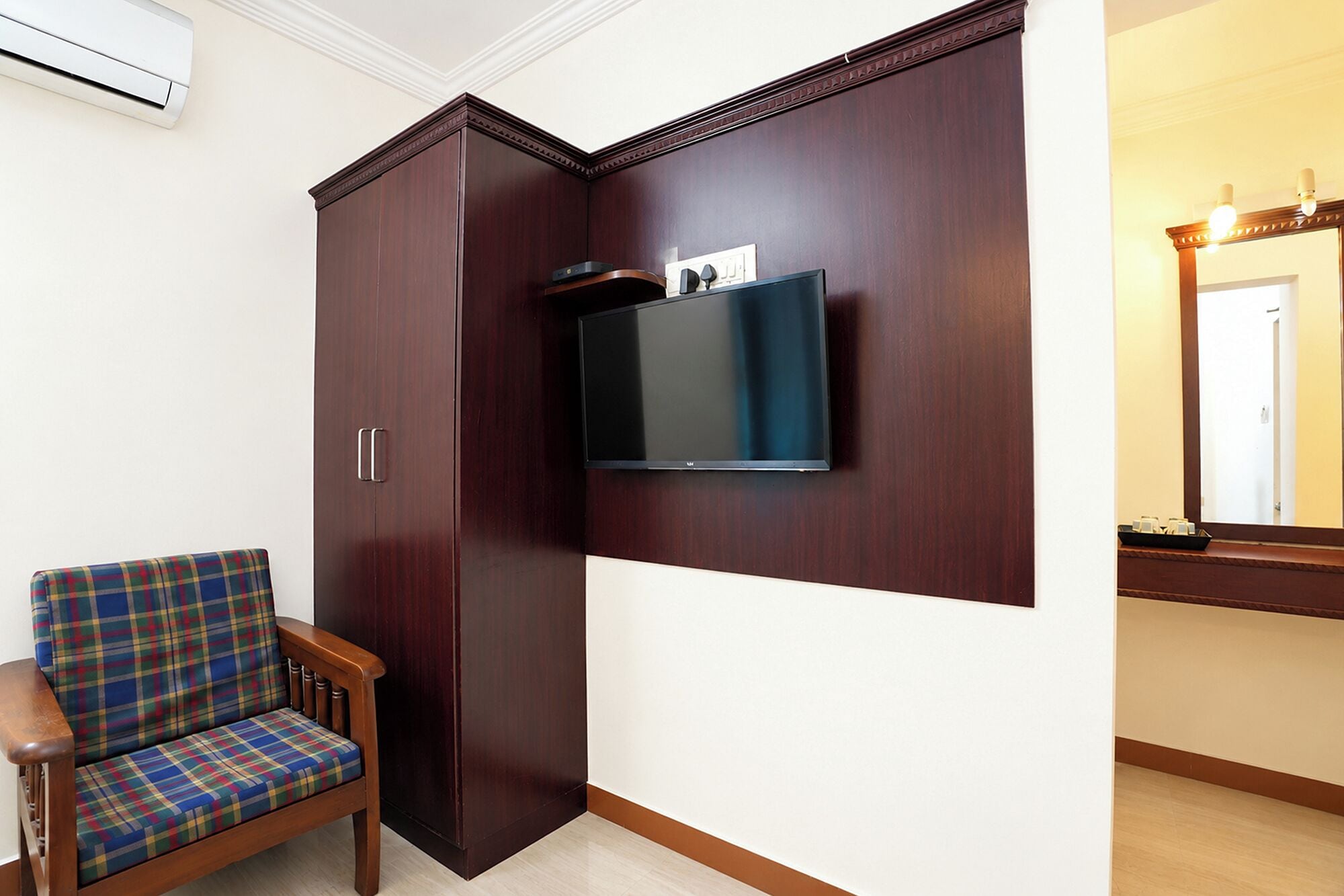 A room with a wardrobe, a television mounted on the wall, and a plaid-patterned chair.