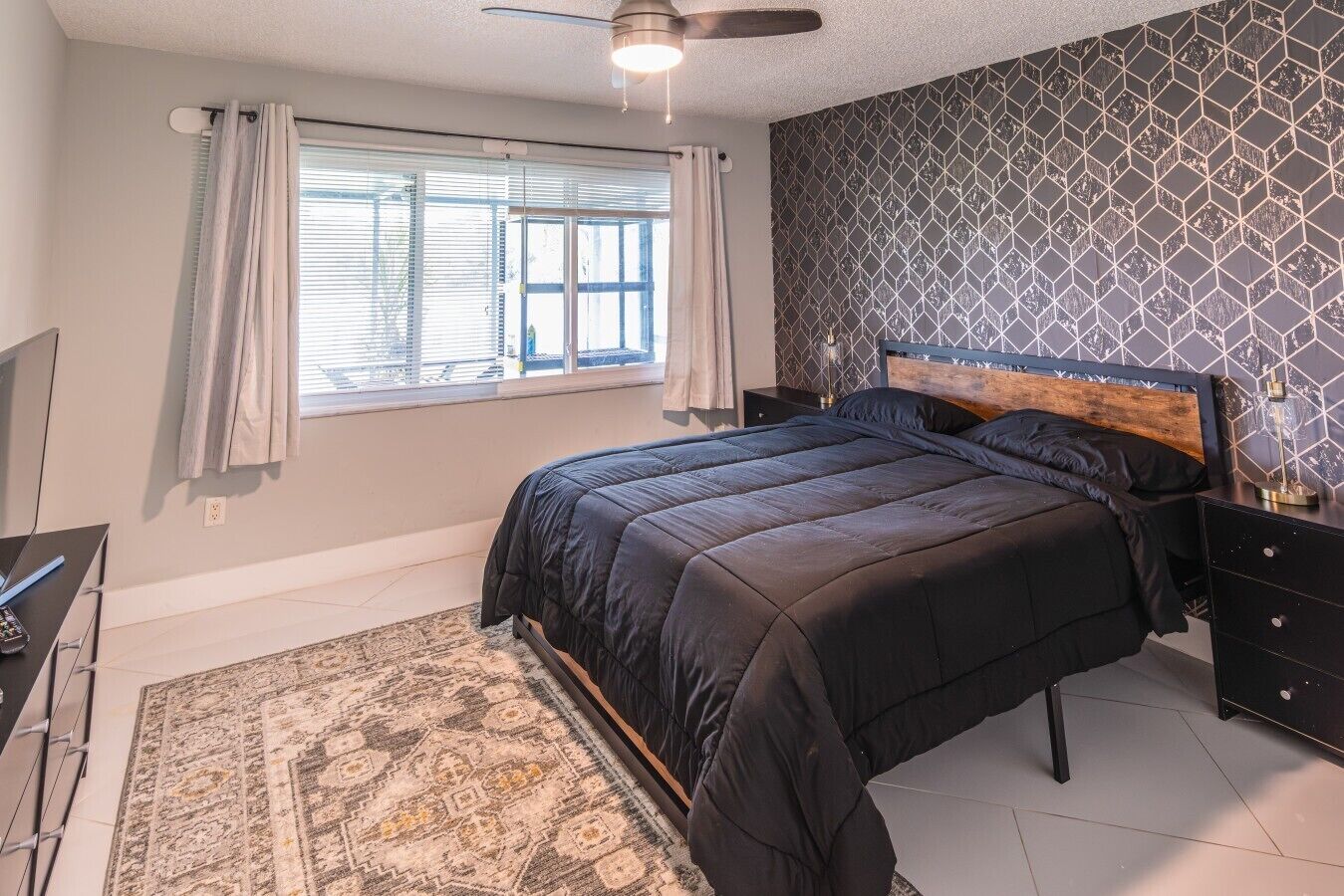 A bedroom with a bed, a nightstand, a desk with a lamp, a window with curtains, and a patterned wall.