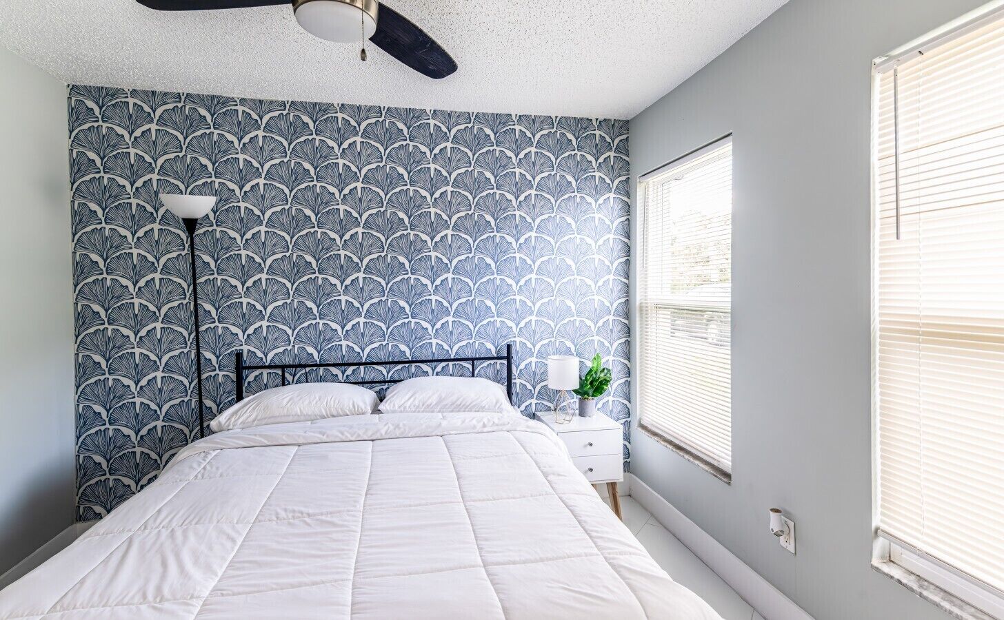 A bedroom with a bed, a ceiling fan, a nightstand with a lamp, and a window with blinds.