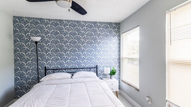 A bedroom with a bed, a ceiling fan, a nightstand with a lamp, and a window with blinds. - Lakeside Oasis Fort Myers (Fort Myers)