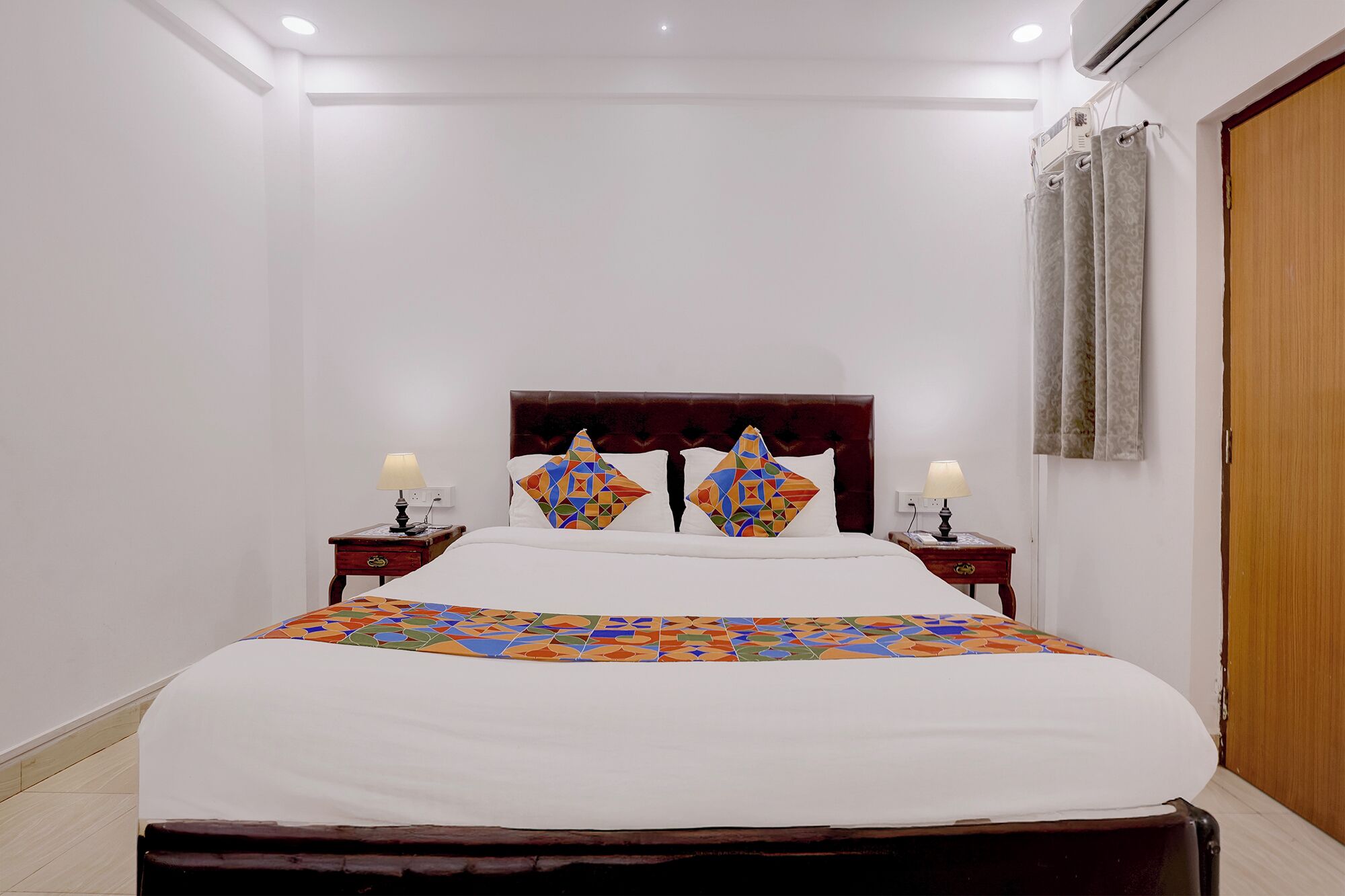 Habitación de hotel con cama, dos mesitas de noche con lámparas, cabecero de madera y puerta de madera.