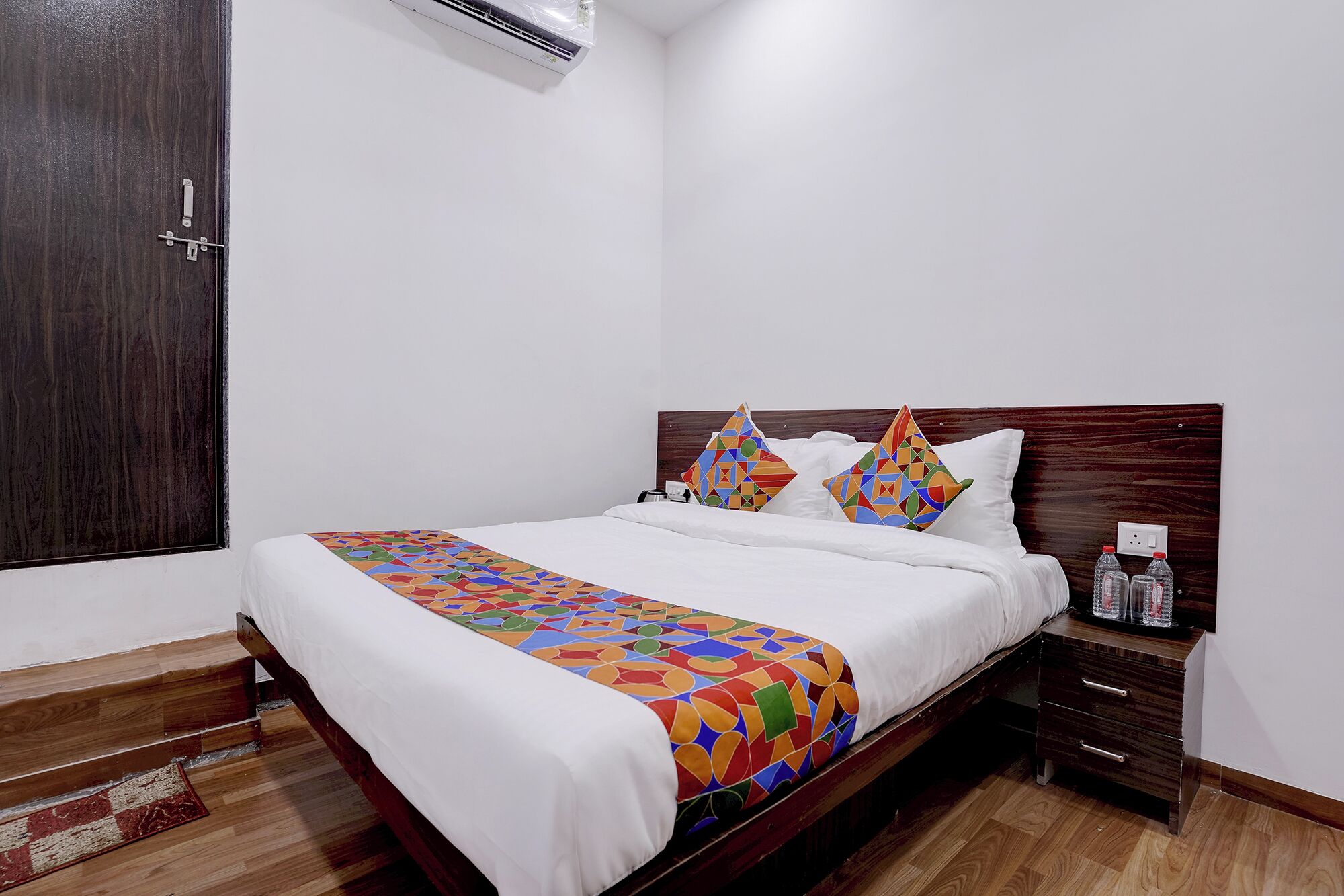 A hotel room with a wooden bed, white bedding, colorful geometric-patterned pillows, a bedside table with bottles, and a wooden floor.