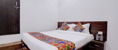 Una habitación de hotel con cama de madera, ropa de cama blanca, almohadas con estampados geométricos de colores y un mesita de noche con botellas, sobre suelo de madera.