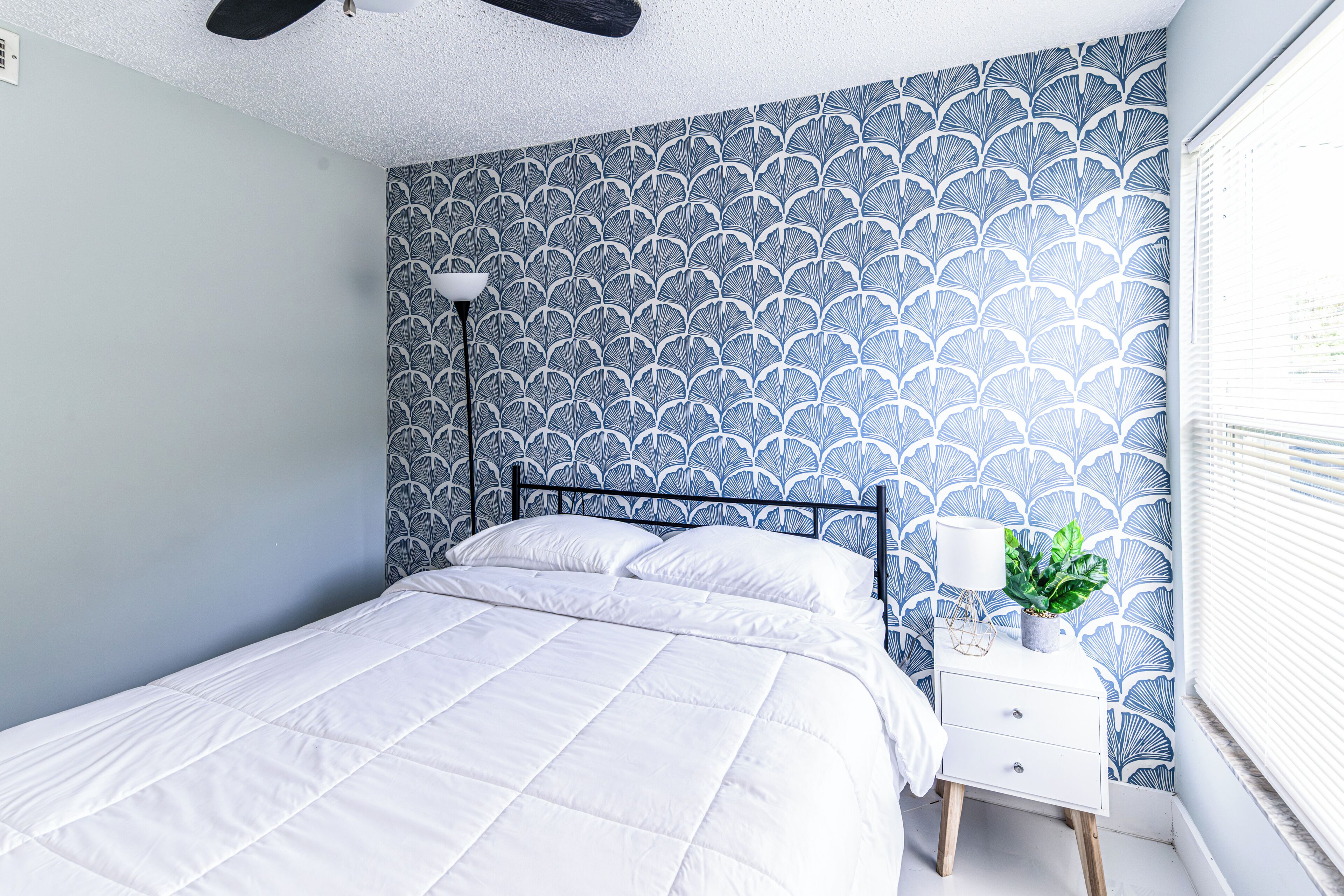 Een slaapkamer met een bed, nachtkastje, lamp en een raam met zonwering.