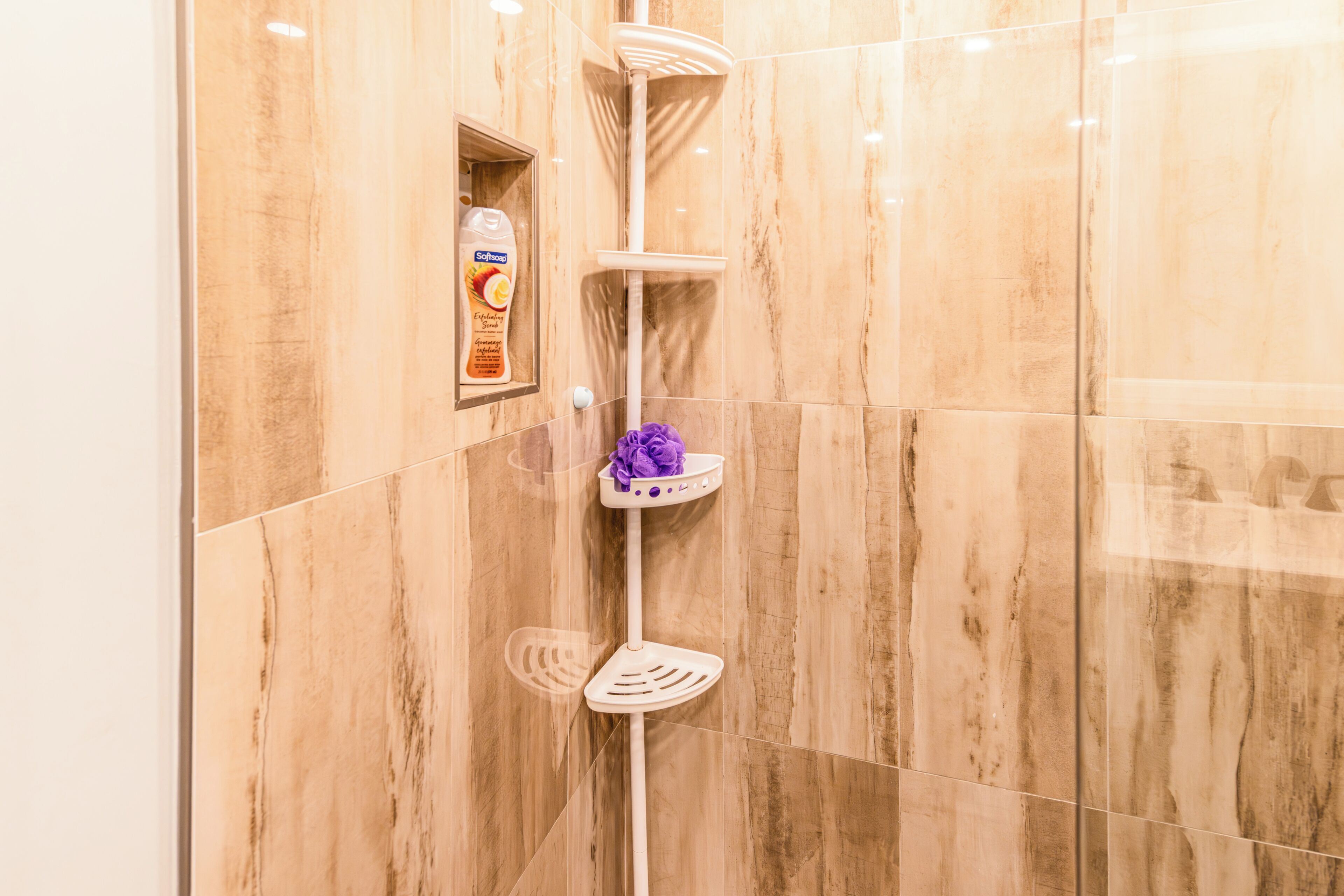 A shower area with a soap dispenser, a purple bath sponge, and a white shelf.
