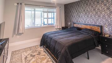 Een slaapkamer met een bed, nachtkastje, bureau, stoel, raam met gordijnen en een plafondventilator.