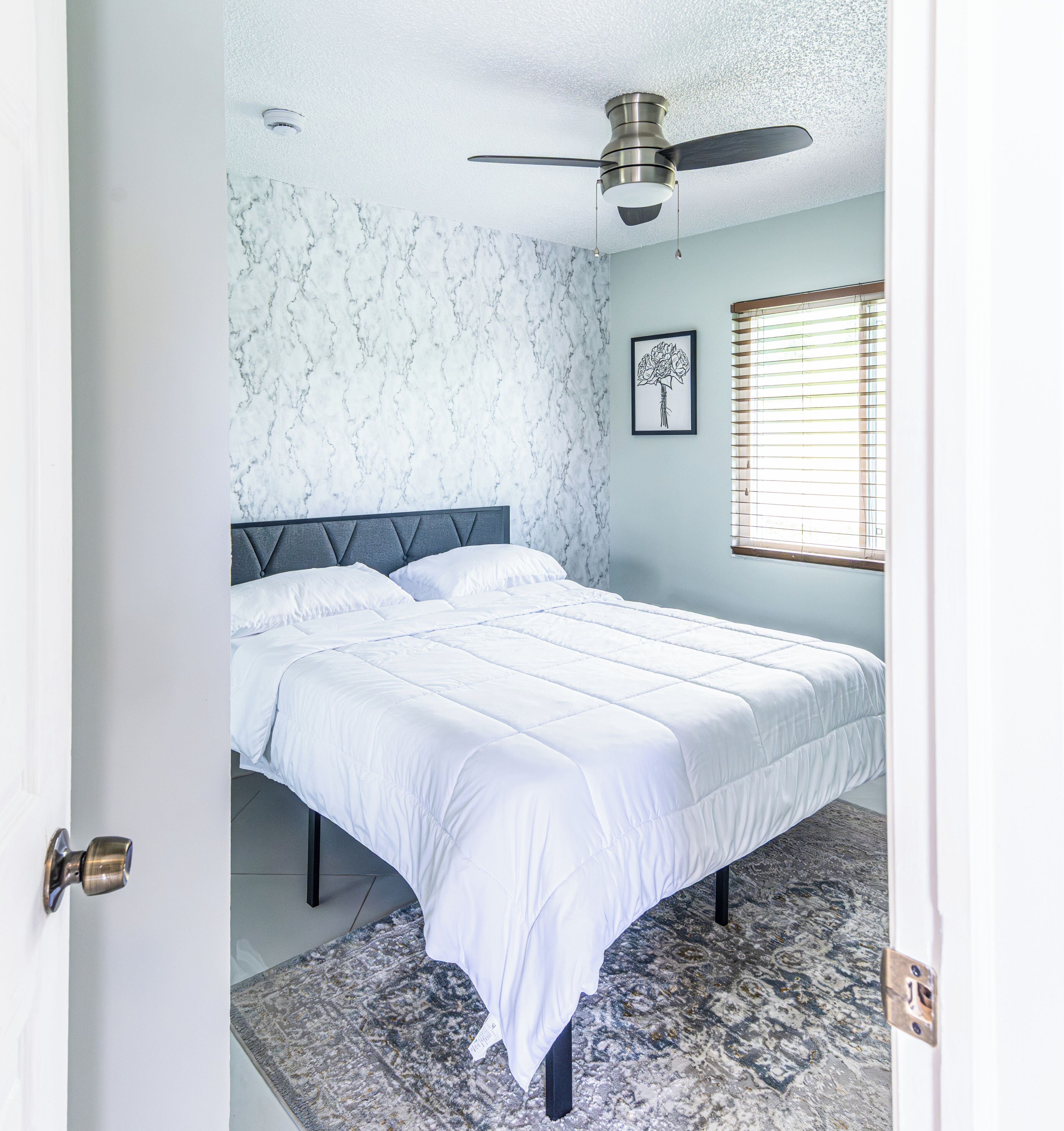 A bedroom with a bed, a ceiling fan, a window with blinds, and a marble-patterned wall.