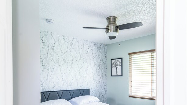 Ein Schlafzimmer mit einem Bett, einem Deckenventilator, einem Fenster mit Jalousien und einer Wand mit Marmoroptik.