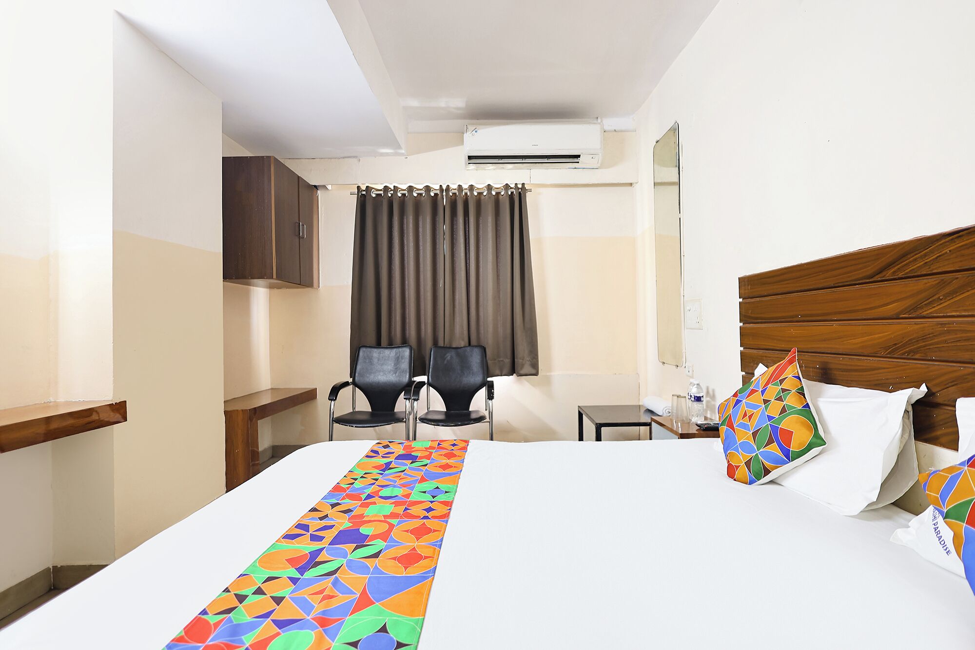Habitación de hotel con cama, escritorio, dos sillas, espejo y aire acondicionado.