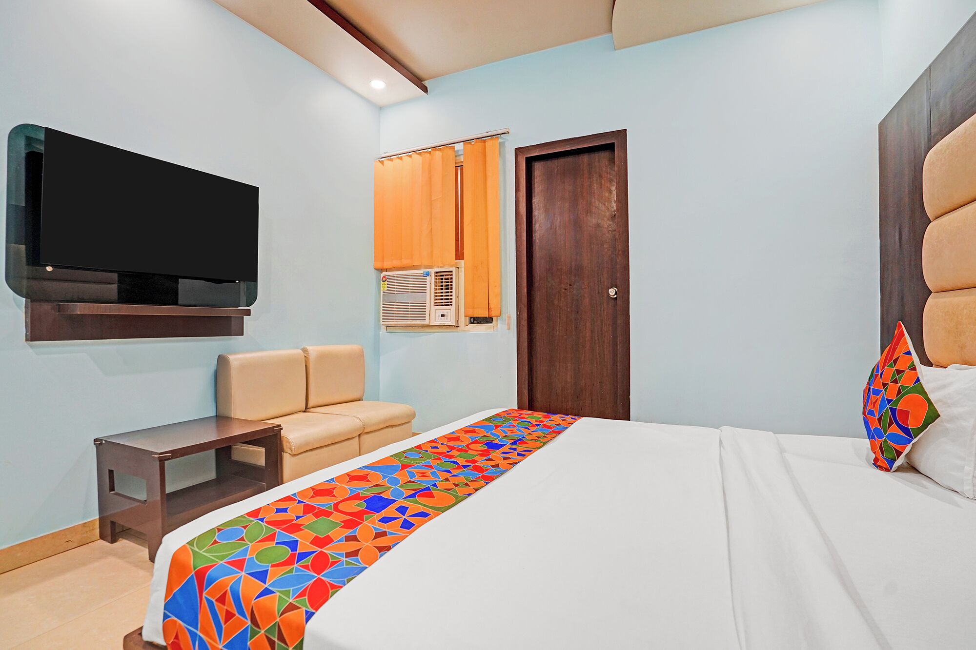 Una habitació d'hotel amb un llit, una televisió penjada a la paret, una taula petita, una cadira i una porta.