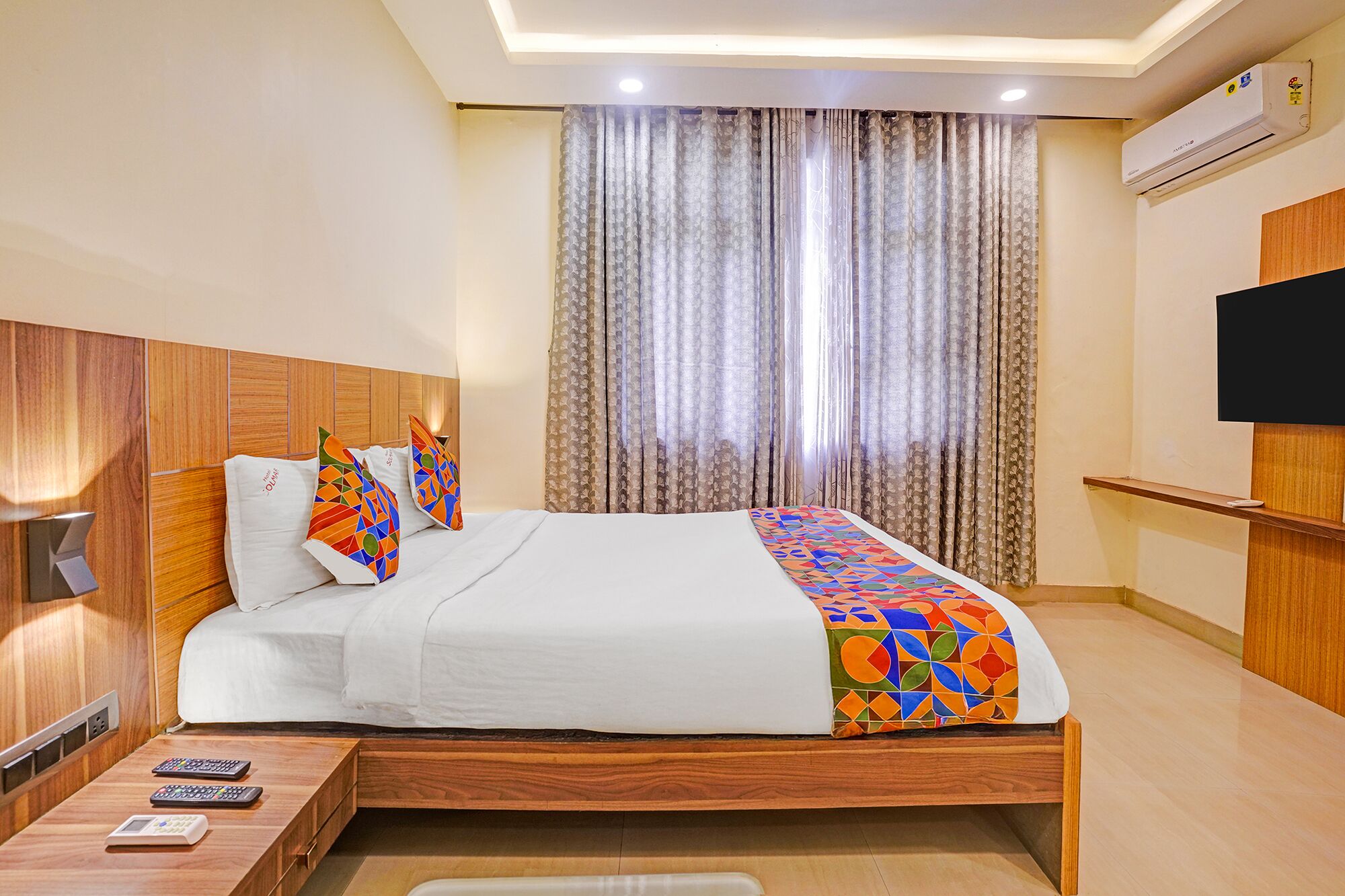 Habitación de hotel con cabecera de madera, cama con ropa de cama blanca y almohadas geométricas de colores, mesita de noche con controles remotos y un gran ventanal con cortinas estampadas.