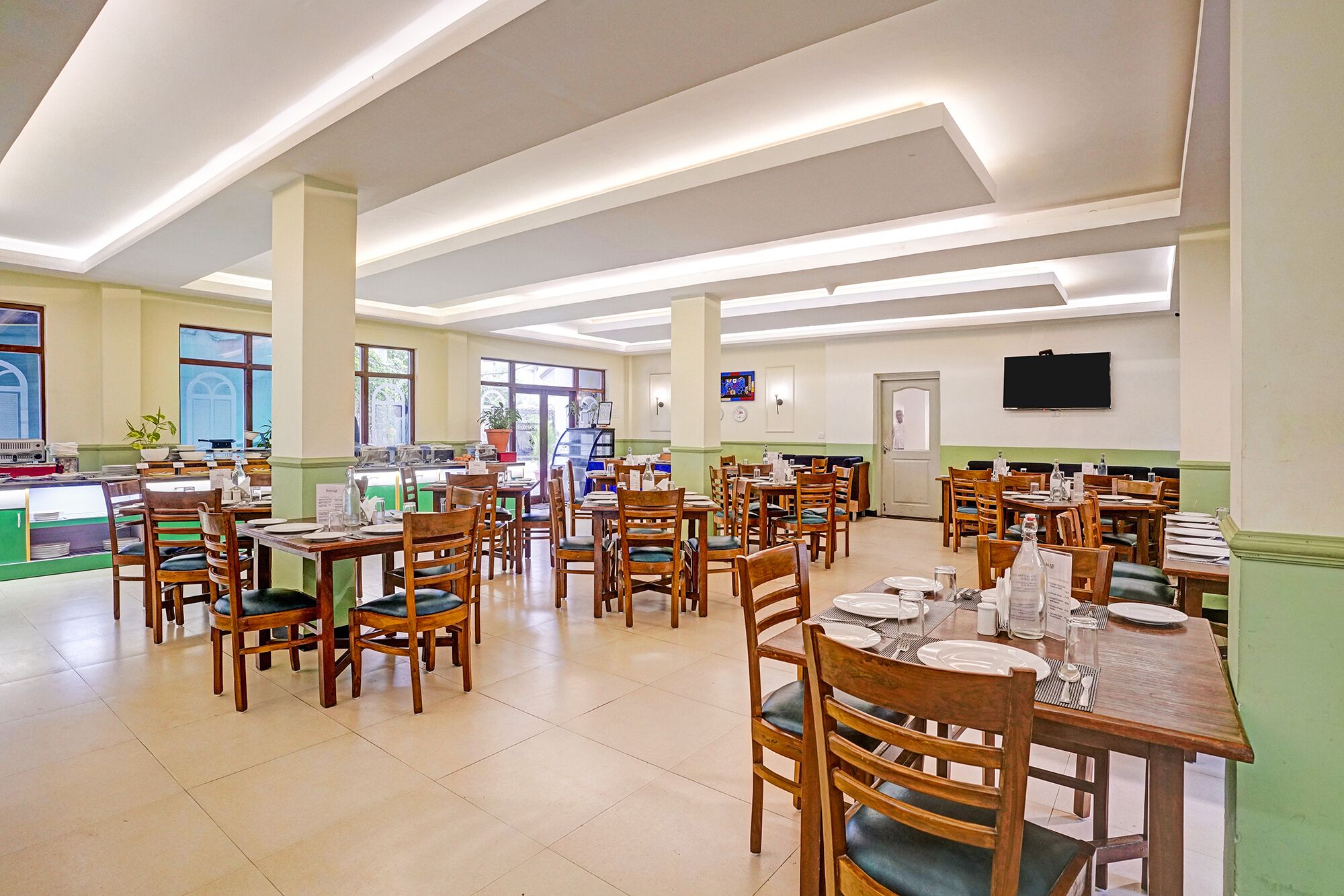 Uma sala de jantar espaçosa com várias mesas e cadeiras, uma televisão na parede e grandes janelas que proporcionam iluminação natural.