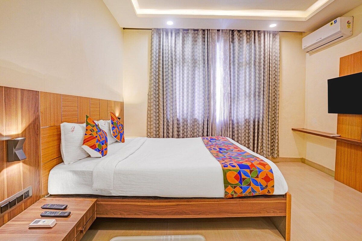 A hotel room with a wooden headboard, a bed with white bedding and colorful geometric pillows, a bedside table with remote controls, and a large window with patterned curtains.