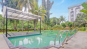 Una piscina con zona cubierta, rodeada de exuberante vegetación y un edificio al fondo.