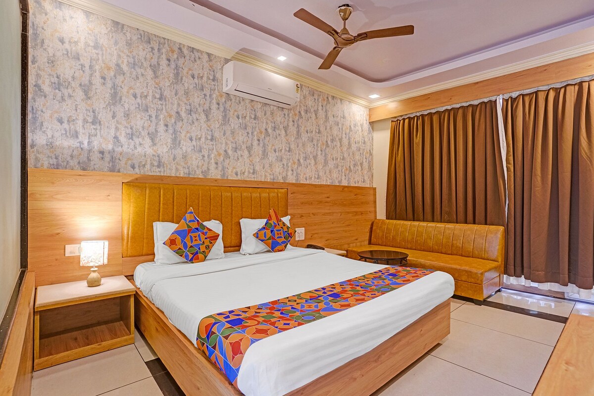 Habitación de hotel con una cama grande, cabecera de madera, ventilador de techo, equipo de aire acondicionado, mesita de noche con lámpara y ventana con cortinas.