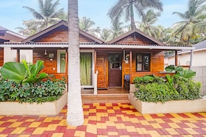 Una casa de madera con entrada de tejas y un porche cubierto, rodeada de exuberante vegetación y palmeras.