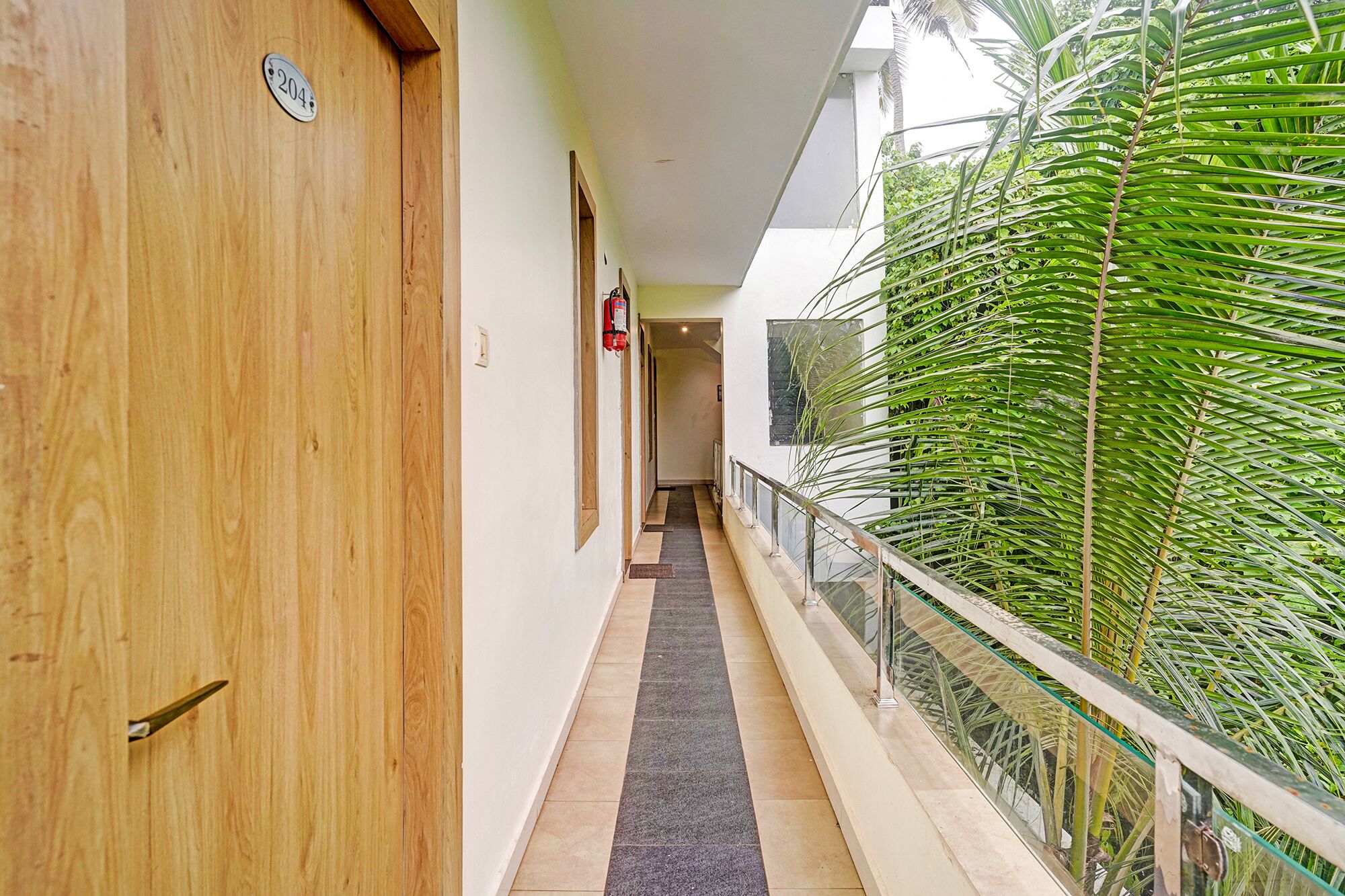 A corridor with a wooden door, a fire extinguisher, and a glass railing overlooking a lush green area.