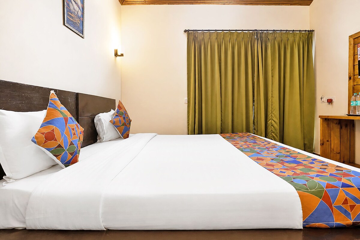 Una habitación de hotel con cama, cabecera, ventana con cortinas y una mesita de madera con botellas.
