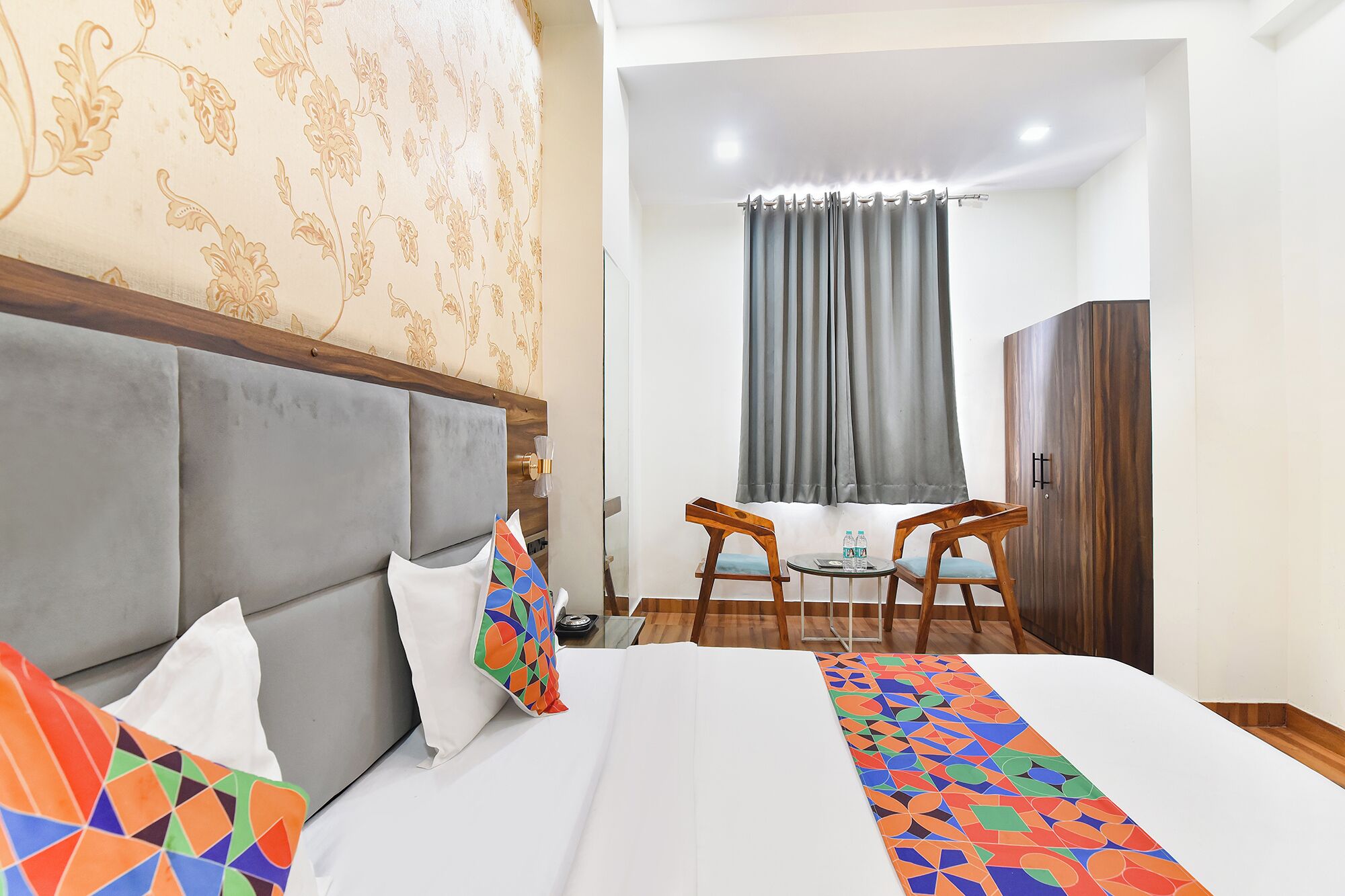 Habitación de hotel con cama, dos sillas, una mesita con botellas de agua, un armario y una ventana con cortinas.