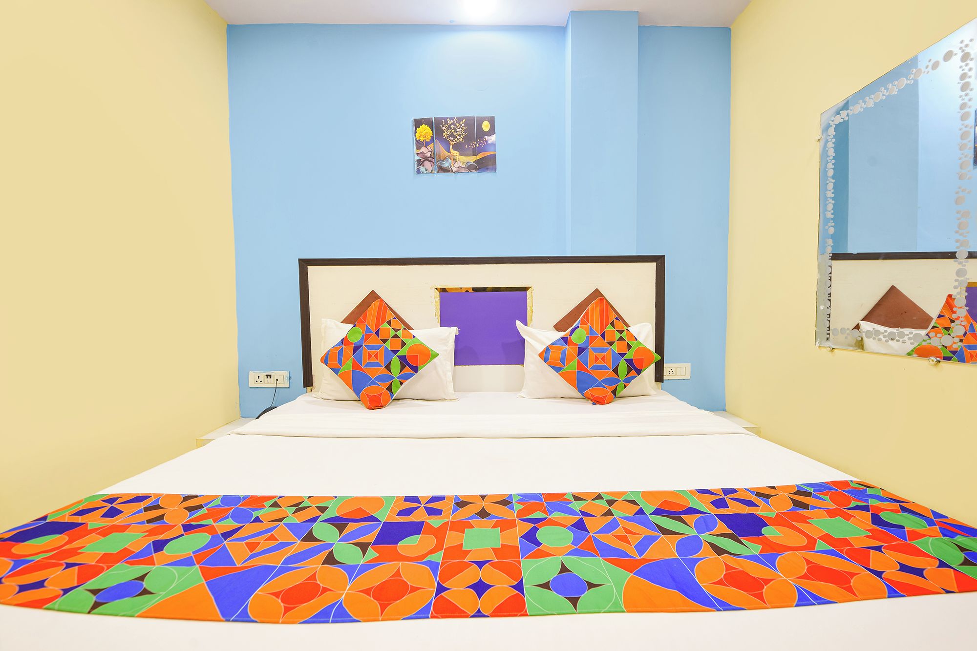 A bed with a colorful geometric-patterned bedspread, a headboard with a mirror, and a small painting on the wall.