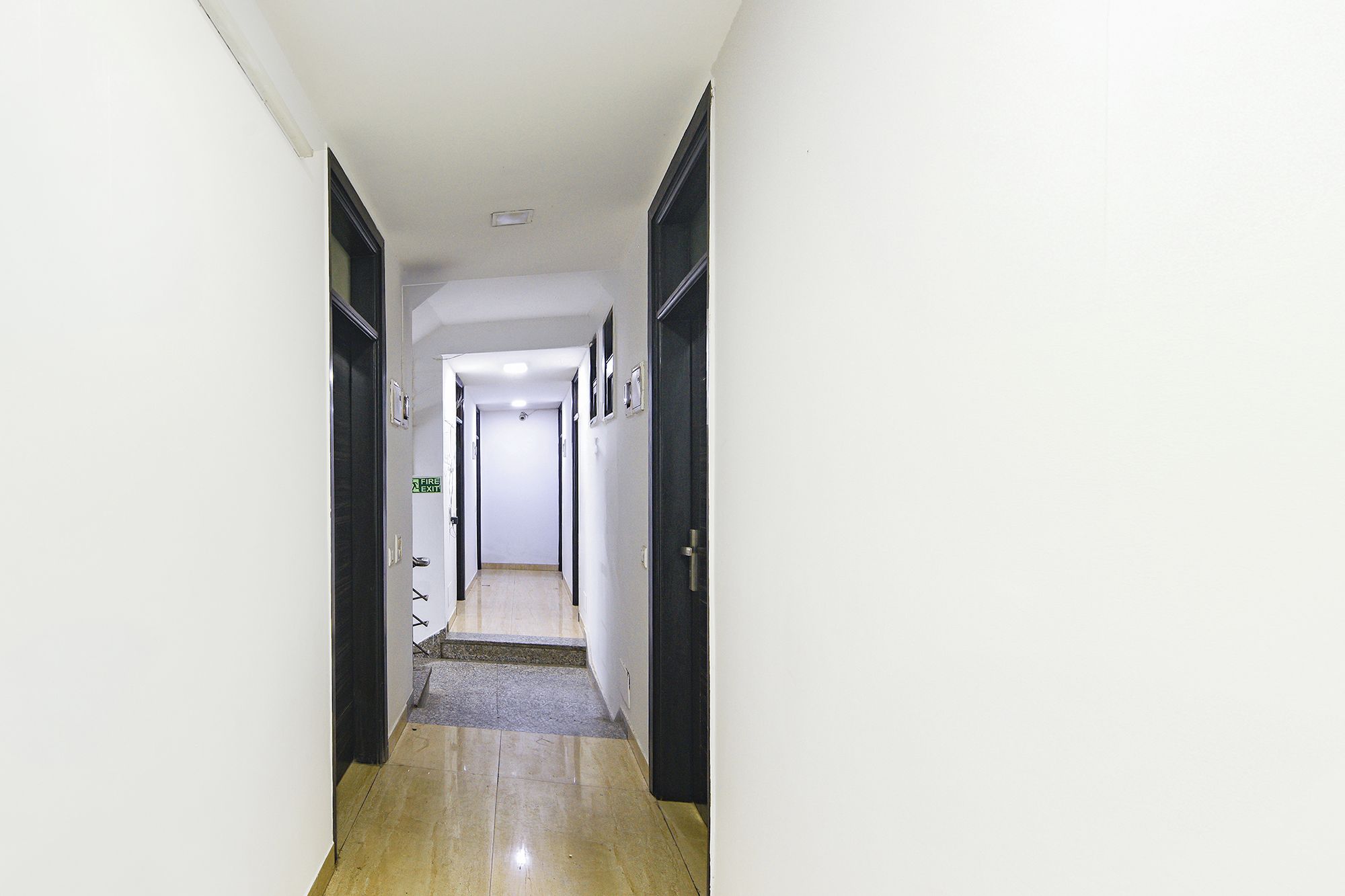 A narrow hallway with a fire exit sign, doors on both sides, and a carpeted floor.