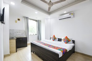 A hotel room with a bed, a ceiling fan, an air conditioning unit, a window with curtains, and a small kitchenette.