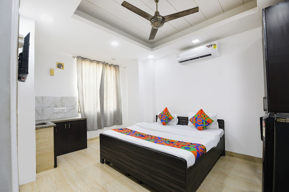 A bedroom with a bed, a ceiling fan, an air conditioning unit, a small kitchenette, and a bathroom with a shower curtain.