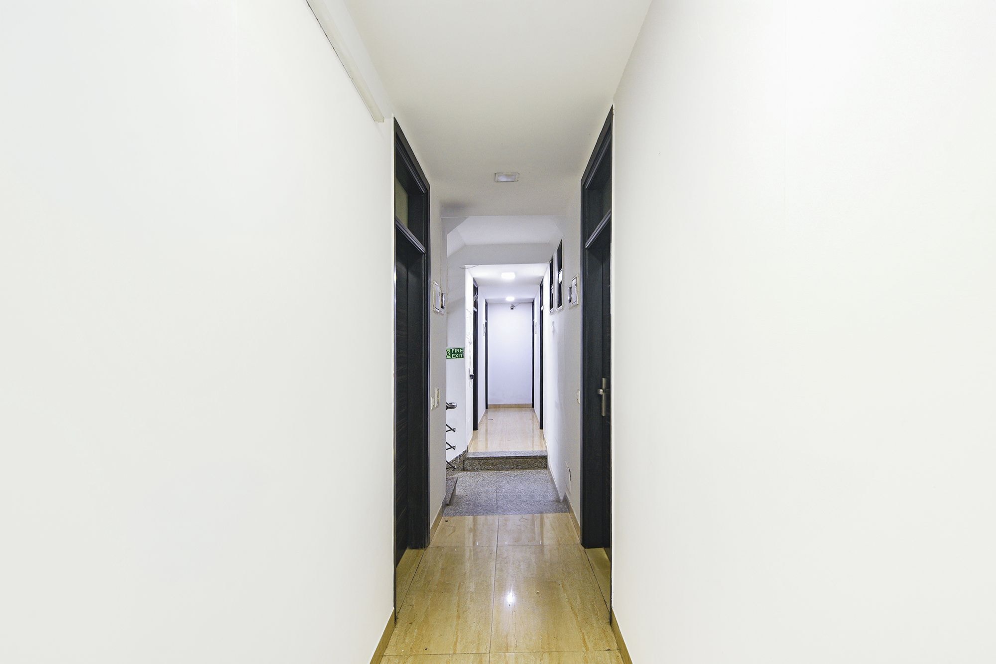 A narrow hallway with a fire exit sign, a door at the end, and a light fixture on the ceiling.