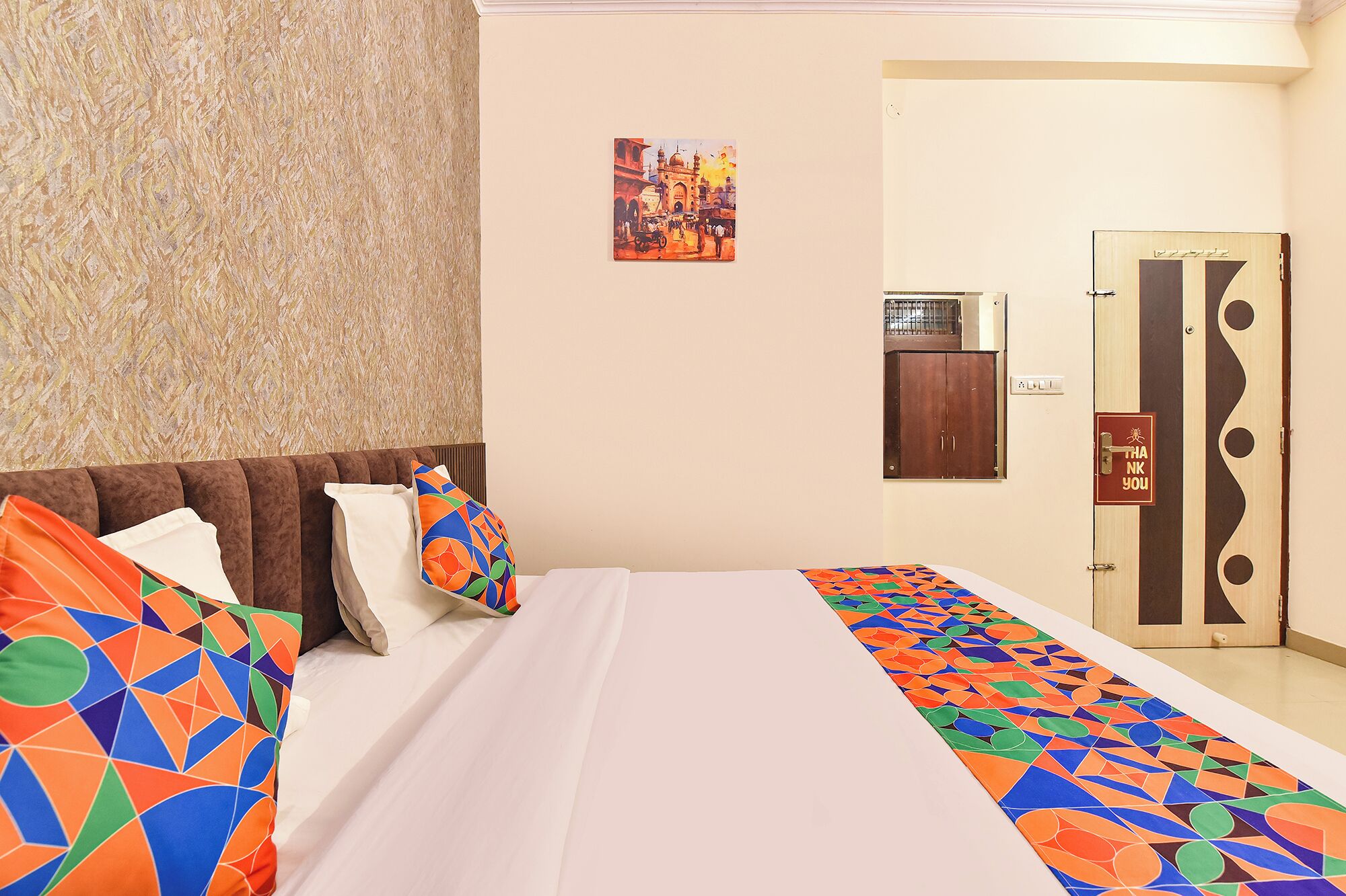 A hotel room with a bed, pillows, a wall-mounted picture, a wardrobe, and a door with a 'Thank You' sign.