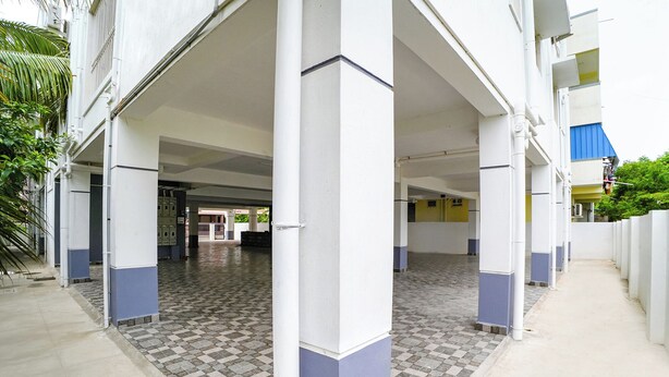 Un espace extérieur abrité, doté d’un sol carrelé et de piliers blancs, qui mène à l’entrée d’un bâtiment.