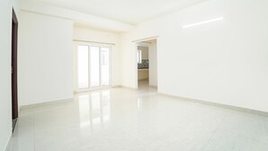 Un espacio amplio y vacío con un gran ventanal corredero que da a un balcón, una entrada a un área de cocina y piso de baldosas.