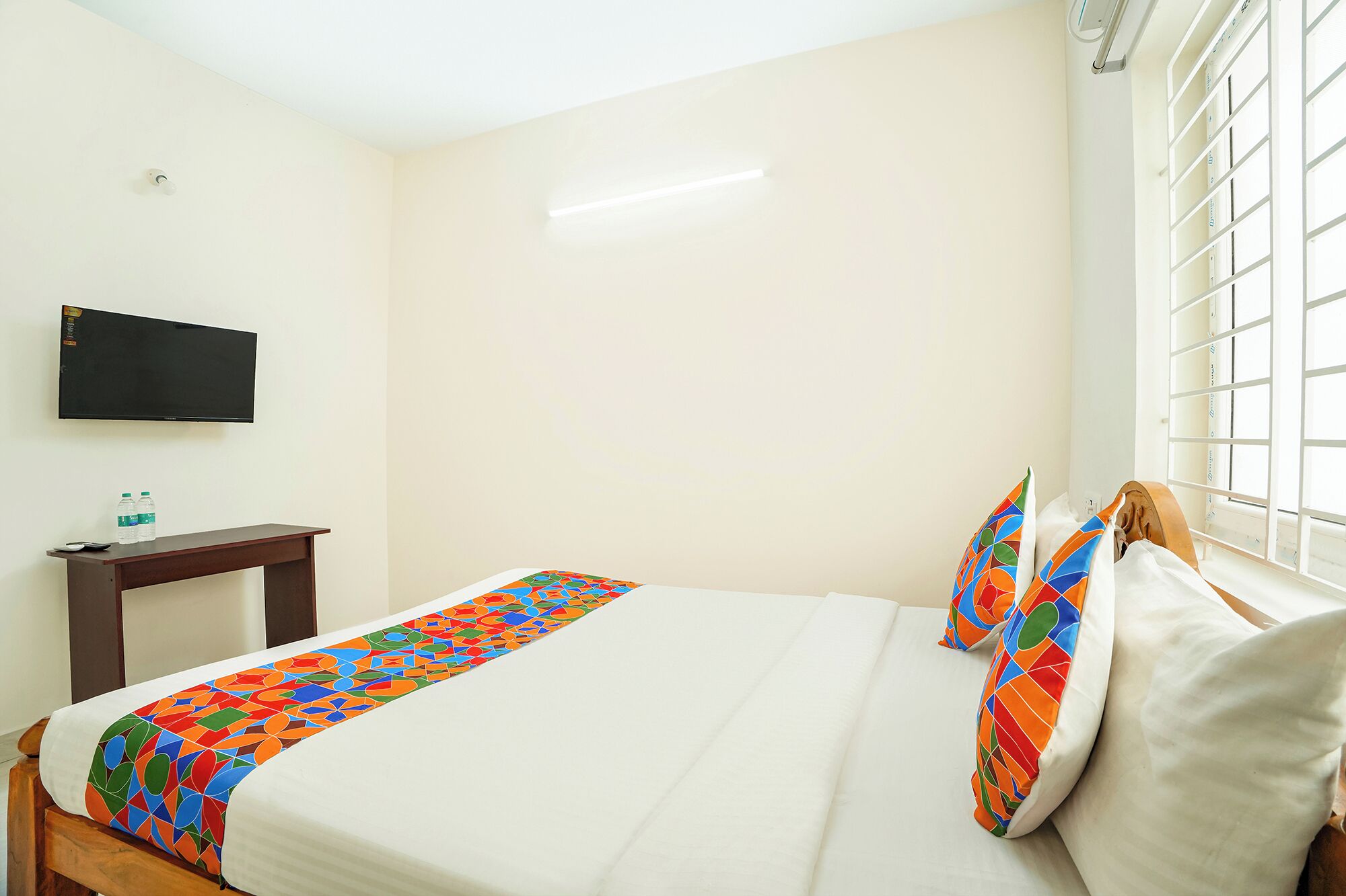 A hotel room with a bed, a TV mounted on the wall, a small table with a remote control, and a window with blinds.