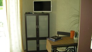 A room with a television on a cabinet, a table with a book and a plant, and a window with curtains. - Little butterfly (Carpentras)