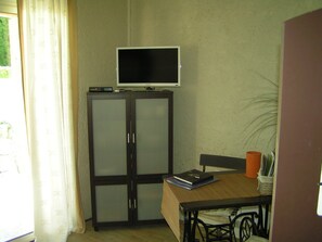 A room with a television on a cabinet, a table with a book and a plant, and a window with curtains.