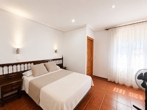 Quarto com cama, mesas de cabeceira, porta de madeira, janela com cortinas e um ventilador.