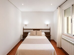 Un dormitori amb un llit de fusta, dues taules de nit, una petita taula i una finestra amb cortines, i un radiador.