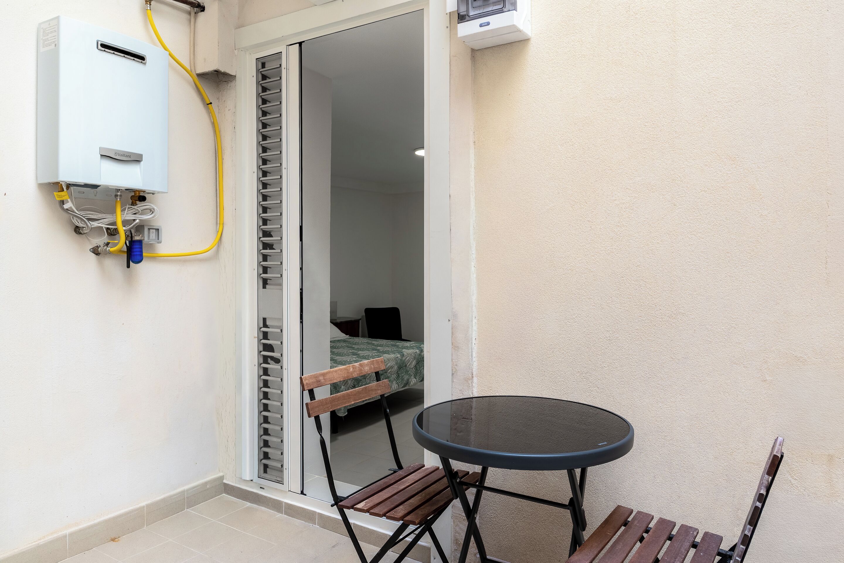 Una habitación con puerta corrediza que da a un balcón, una pequeña mesa redonda con un banco y un calentador de agua instalado en la pared.