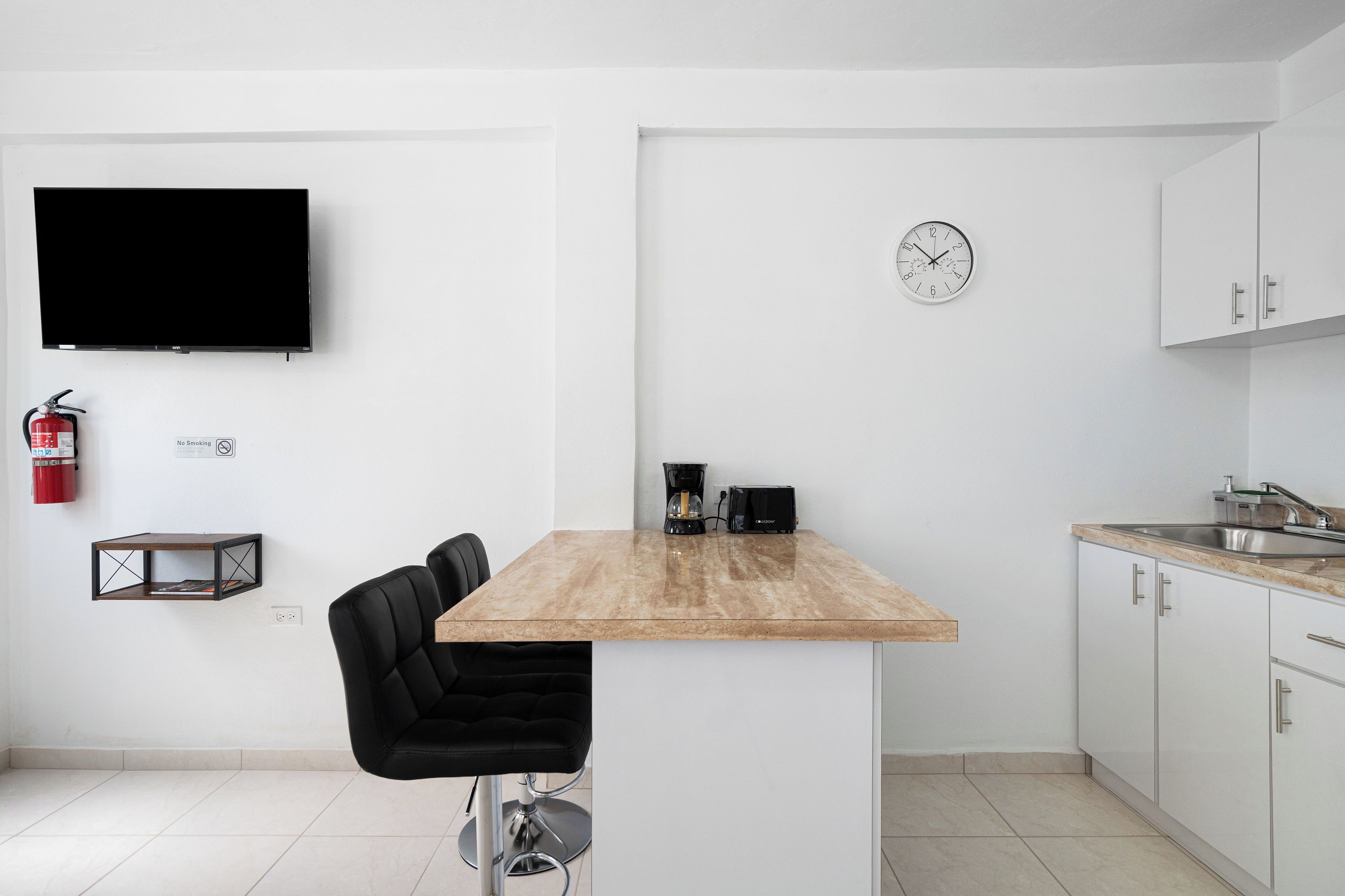 A modern kitchen with a wooden table, black chairs, a coffee machine, a clock, and a flat-screen TV.