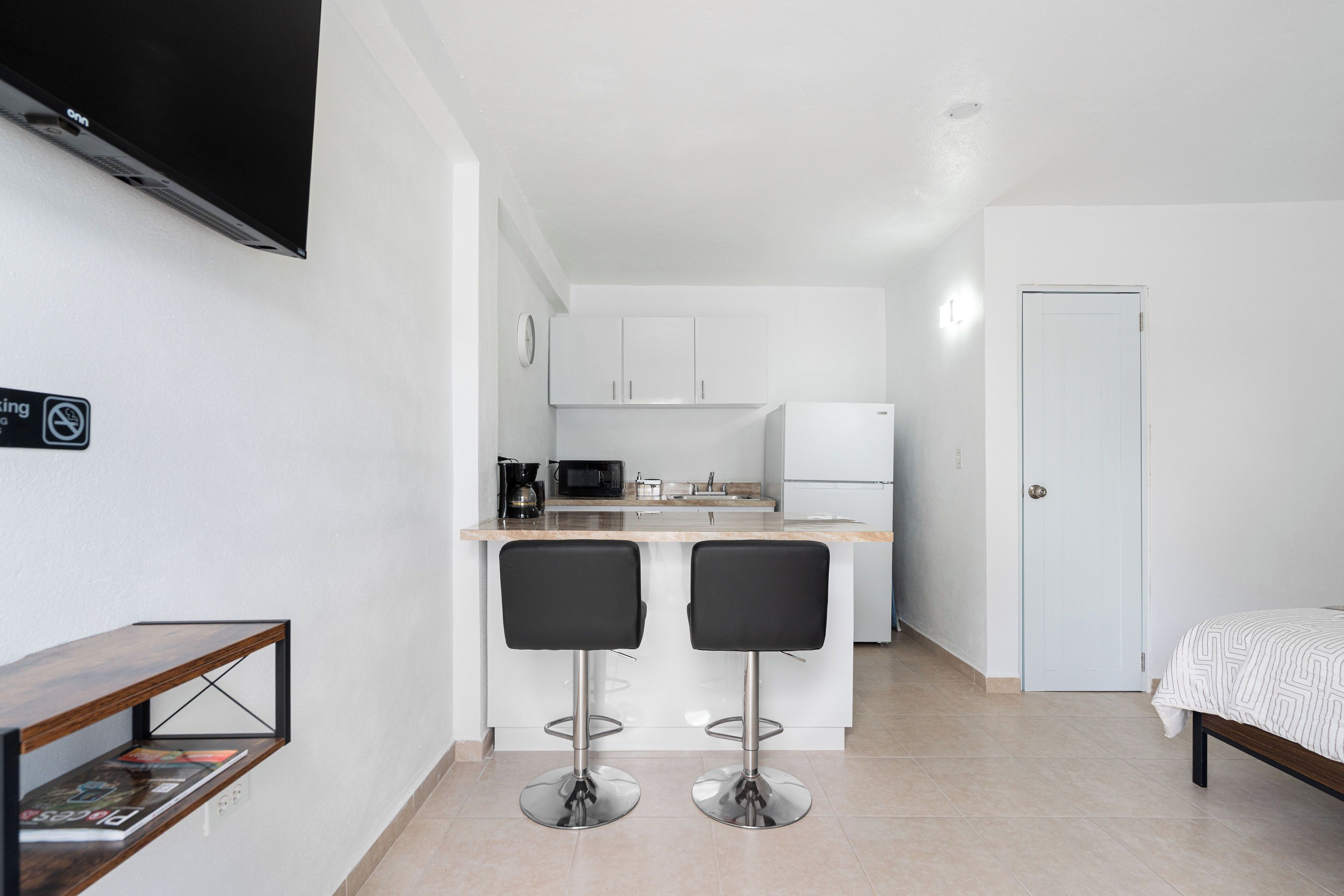 A compact kitchen with a microwave, coffee maker, and refrigerator. There are two bar stools and a small dining table. A bed with a patterned blanket is visible in the adjacent room.