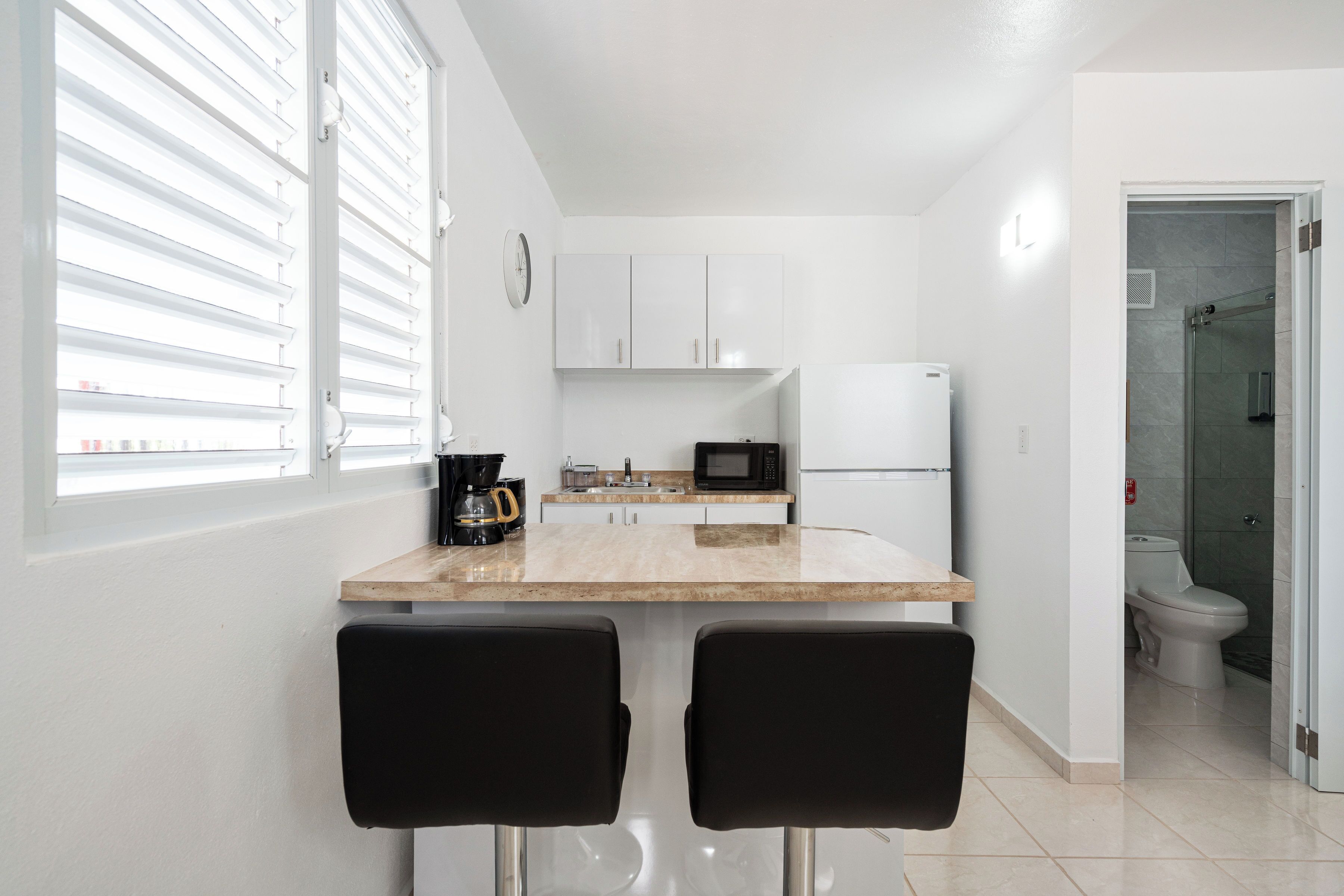Una cocina compacta con una encimera, dos sillas negras, una cafetera, un microondas y un refrigerador.
