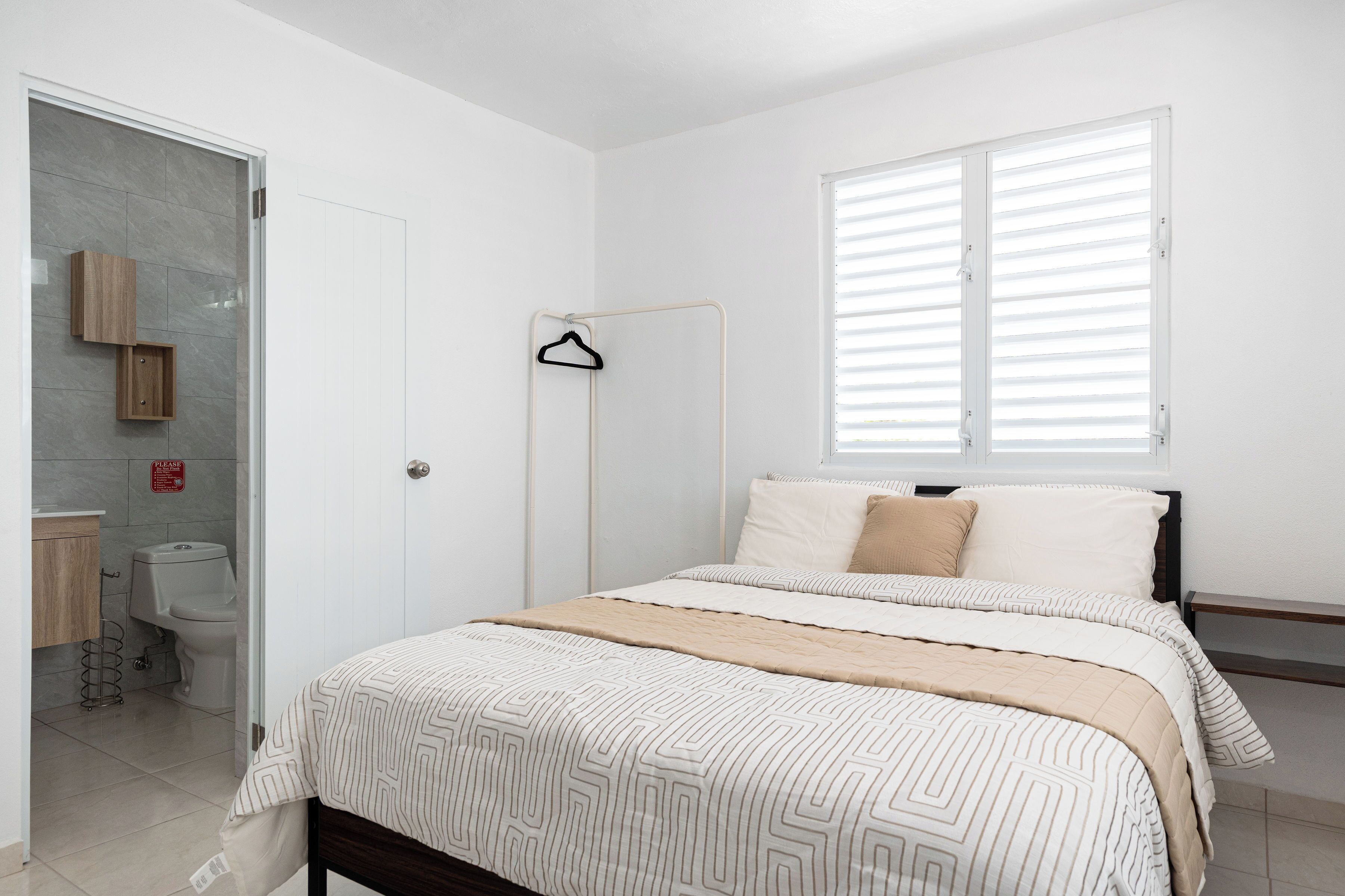 Une chambre avec un lit, une fenêtre avec des stores, un portant à vêtements et des toilettes.