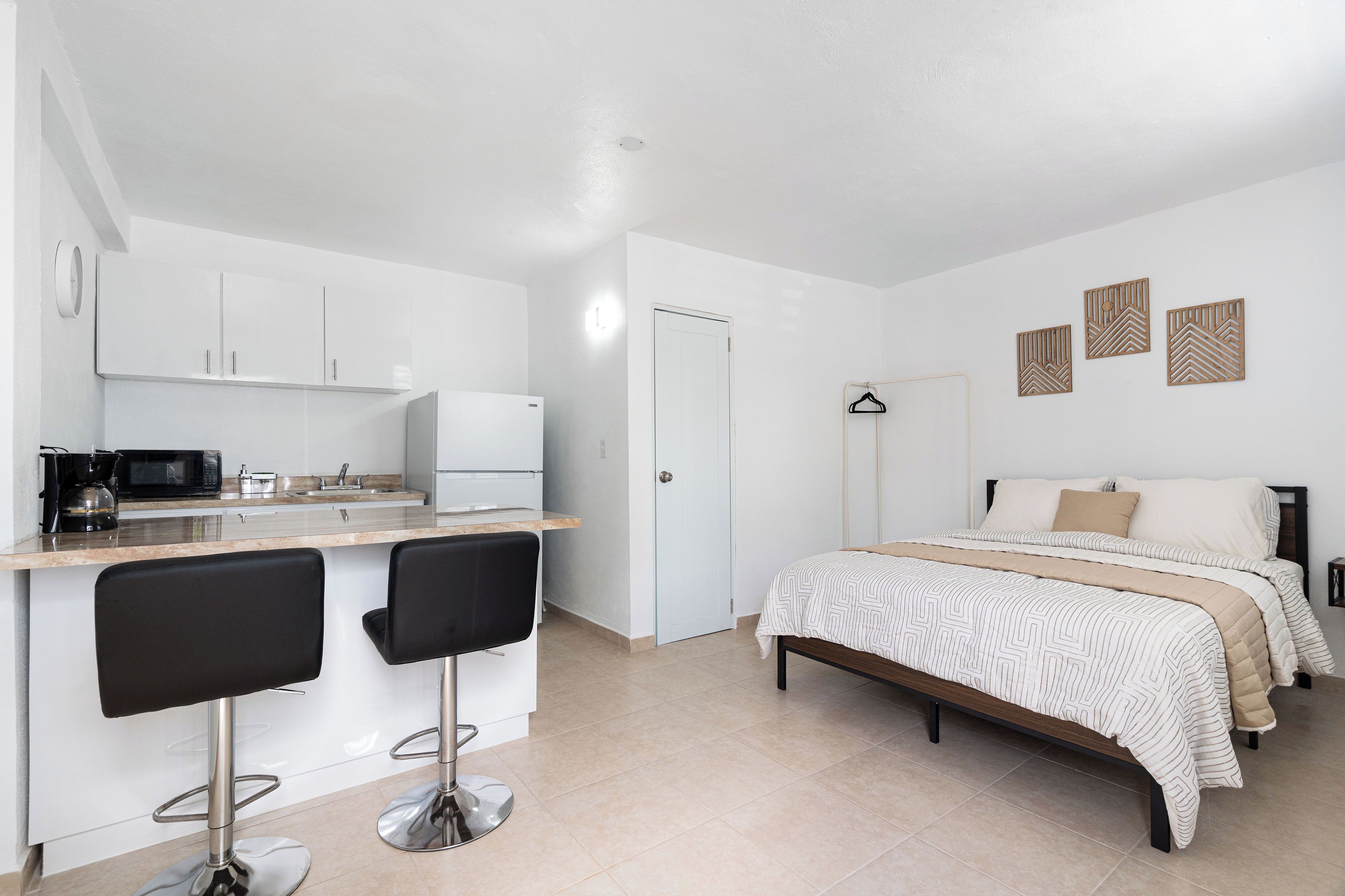 Ein kompaktes Zimmer mit einer Küchenzeile, einem Bett mit gemusterten Betttextilien und einer kleinen Sitzecke mit zwei Stühlen.